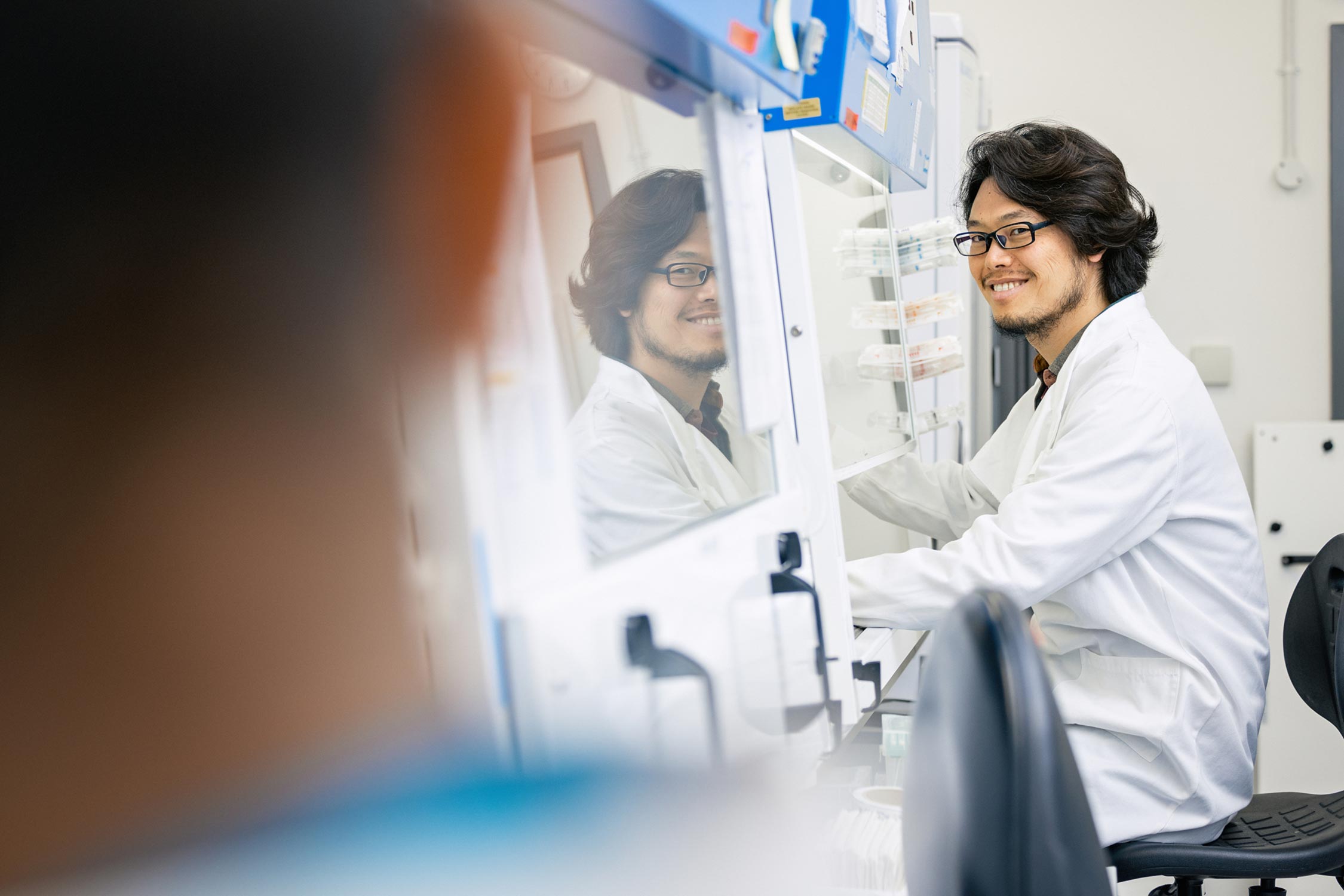 A researcher working in a tissue culture hood looks towards a colleague in the foreground